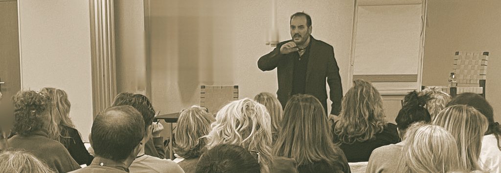 El profesor explica de pie ante un grupo de alumnos en una clase presencial del Instituto Área Zentro, vista desde el fondo de la sala